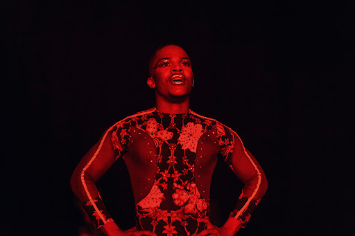 A man with hands on hips is illuminated by red lighting on a dark stage wearing a black and red patterned body suit.