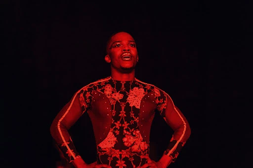 A man with hands on hips is illuminated by red lighting on a dark stage wearing a black and red patterned body suit.