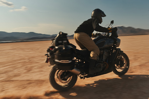 A person rides a motorcycle across a desert landscape on a sunny day, kicking up dust. The motorcycle is equipped with storage bags.