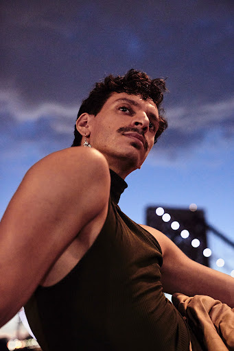 Portrait of a man with dark hair and a moustache wearing a black top, posing outdoors with a city bridge and cloudy sky in the background.