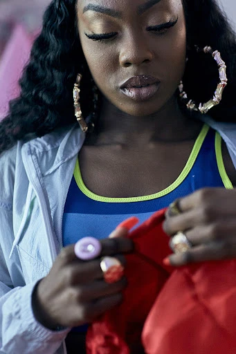 Close-up of stylish African-American woman with long curly hair, hoop earrings, vibrant blue and green top, and a light jacket indoors.