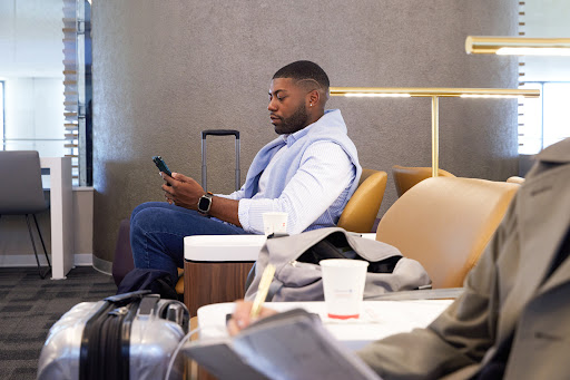 Man in jeans sits in airport lounge, looking at phone, with a light blue sweater draped over shoulders, and a rolling suitcase nearby.