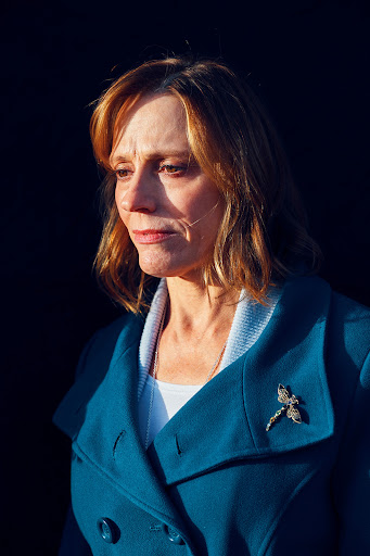 Portrait of a middle-aged woman with light brown hair wearing a teal coat, standing in front of a dark, shadowed background.