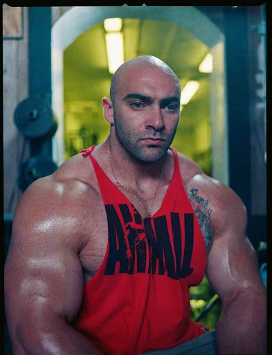 Muscular bald man in a red tank top poses in an indoor gym setting, showcasing his physique with equipment visible in the background.