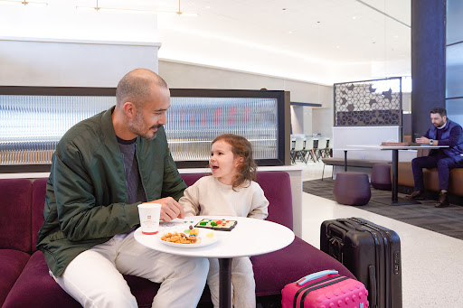 Father and daughter at a cafe table with snacks and a drawing, sitting on a purple couch, suitcases nearby, enjoying time together.