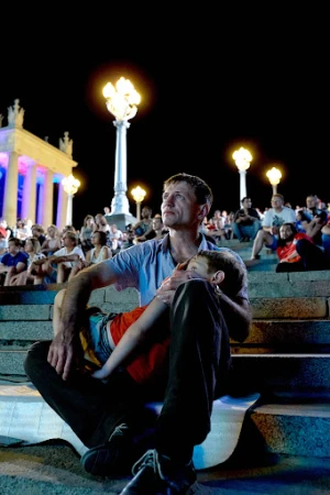 Father and child sit on steps at night. Man looks up while embracing sleeping child on his lap in outdoor urban setting with crowd.