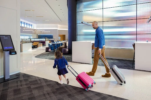 Father and child pulling suitcases in a modern airport lounge with seating, large windows, and a self-service kiosk.