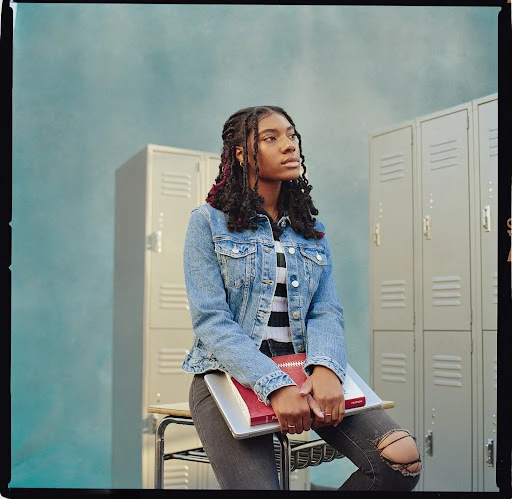 Teenage African-descent student sitting on a desk holding books in front of lockers wearing a denim jacket and ripped jeans in school.