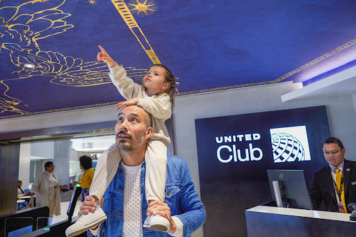 Father with young daughter on shoulders, pointing up at ceiling art in United Club lounge, airport travel experience.