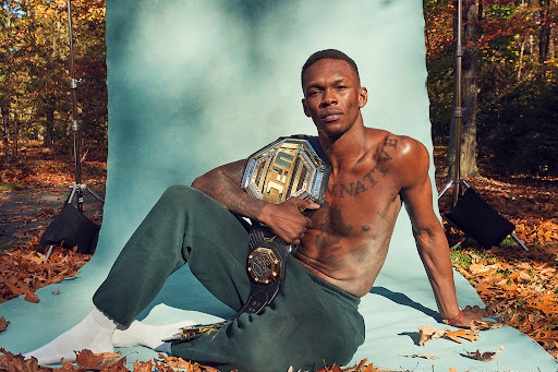 Shirtless fighter Izzy Adesanya poses with UFC championship belt against turquoise backdrop in autumn leaves and trees.