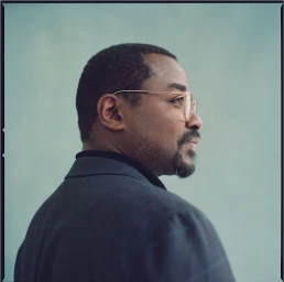 Profile portrait of a Black man with glasses and a goatee wearing a dark jacket against a pale background