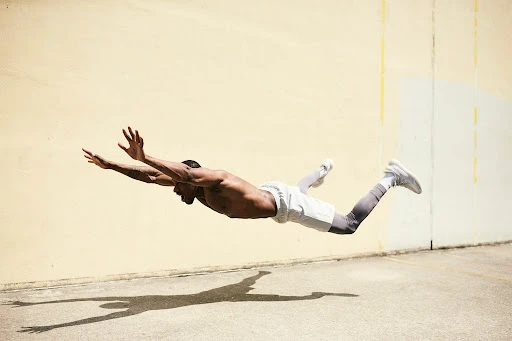 Shirtless man captured mid-air in a horizontal dive above a concrete ground in front of a weathered wall