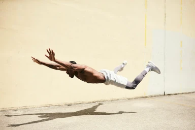 Shirtless man captured mid-air in a horizontal dive above a concrete ground in front of a weathered wall