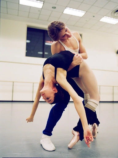 Two ballet dancers performing a dramatic backbend pose inside a bright dance studio with mirrors and ballet barres.