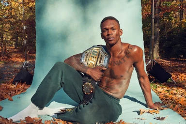 Israel Adesanya seated outdoors holding a UFC championship belt in front of a light blue backdrop on a leaf-covered ground.