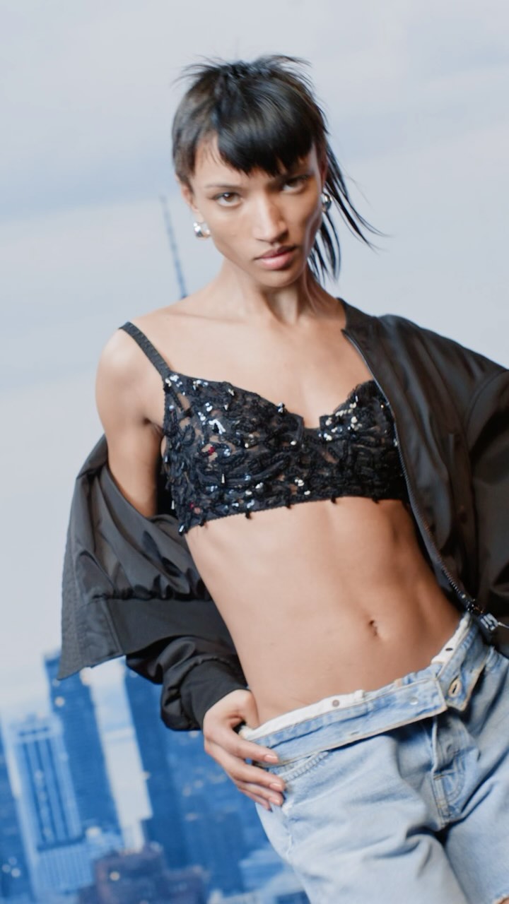 Fashion shot of a woman wearing a black bra top, light blue jeans, and a black jacket against a cityscape background.