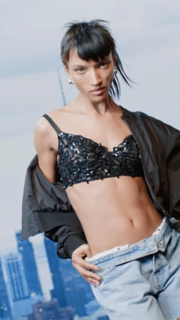Fashion shot of a woman wearing a black bra top, light blue jeans, and a black jacket against a cityscape background.