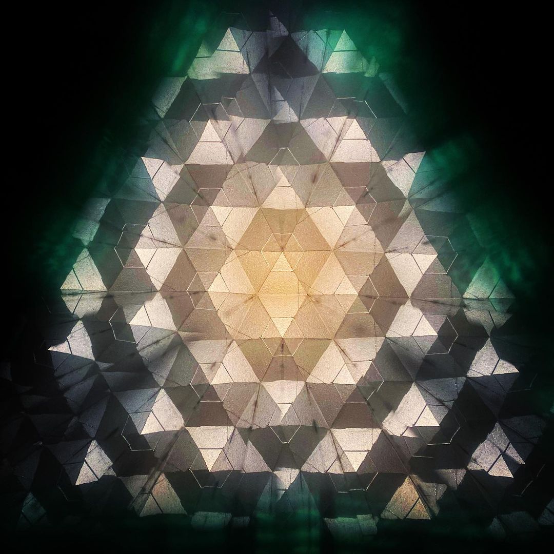 Abstract geometric pattern with triangle shapes and a central glow, creating a kaleidoscope-like effect in shades of green, beige, and black.
