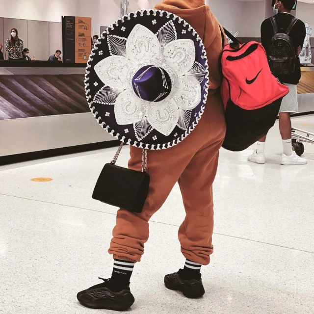 Person in sweats and sneakers carrying a large decorative sombrero and bag, standing on a light-colored floor in an airport or station.