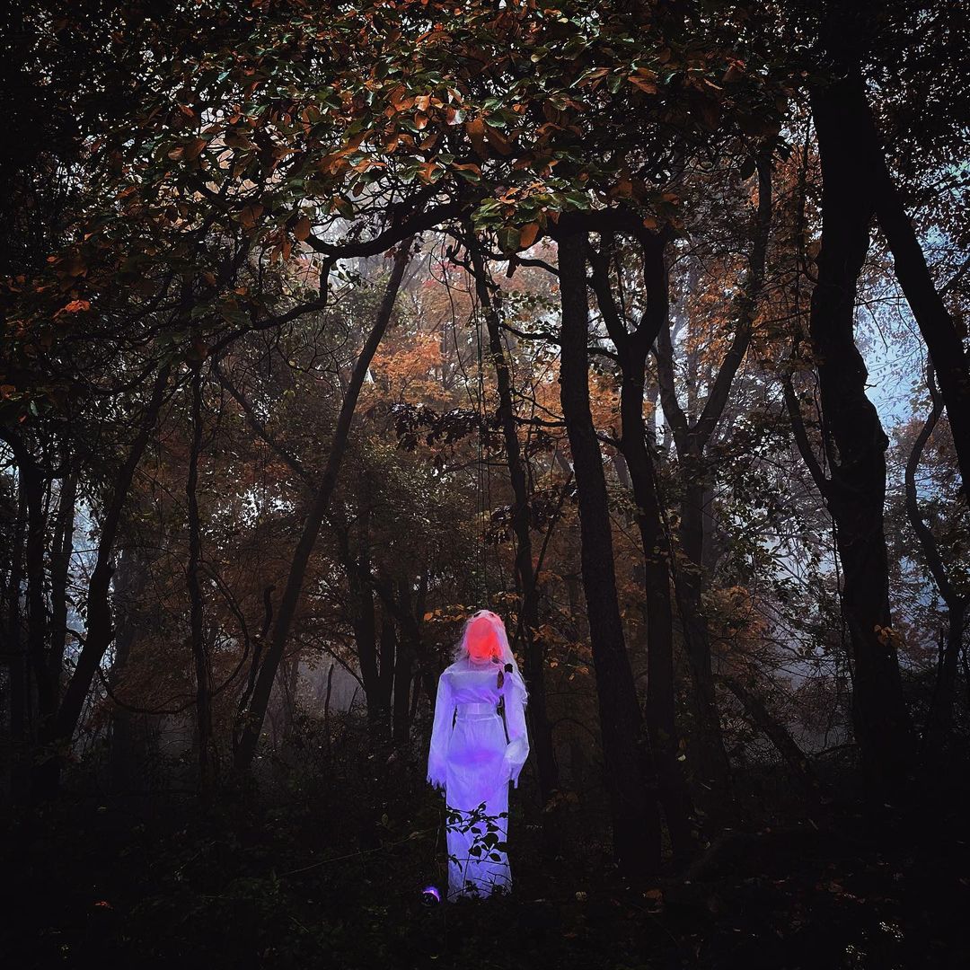 Person in white outfit with glowing head standing in a foggy forest at night, creating an eerie and mysterious Halloween scene.