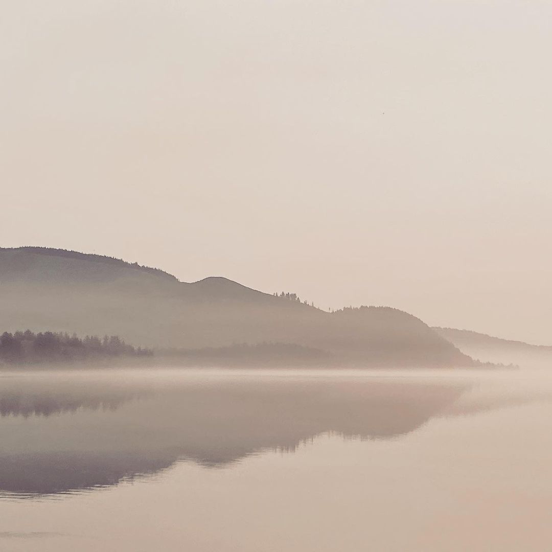 Misty lake landscape with mountains in the background reflecting in the water creating a tranquil, serene scene at dusk or dawn.