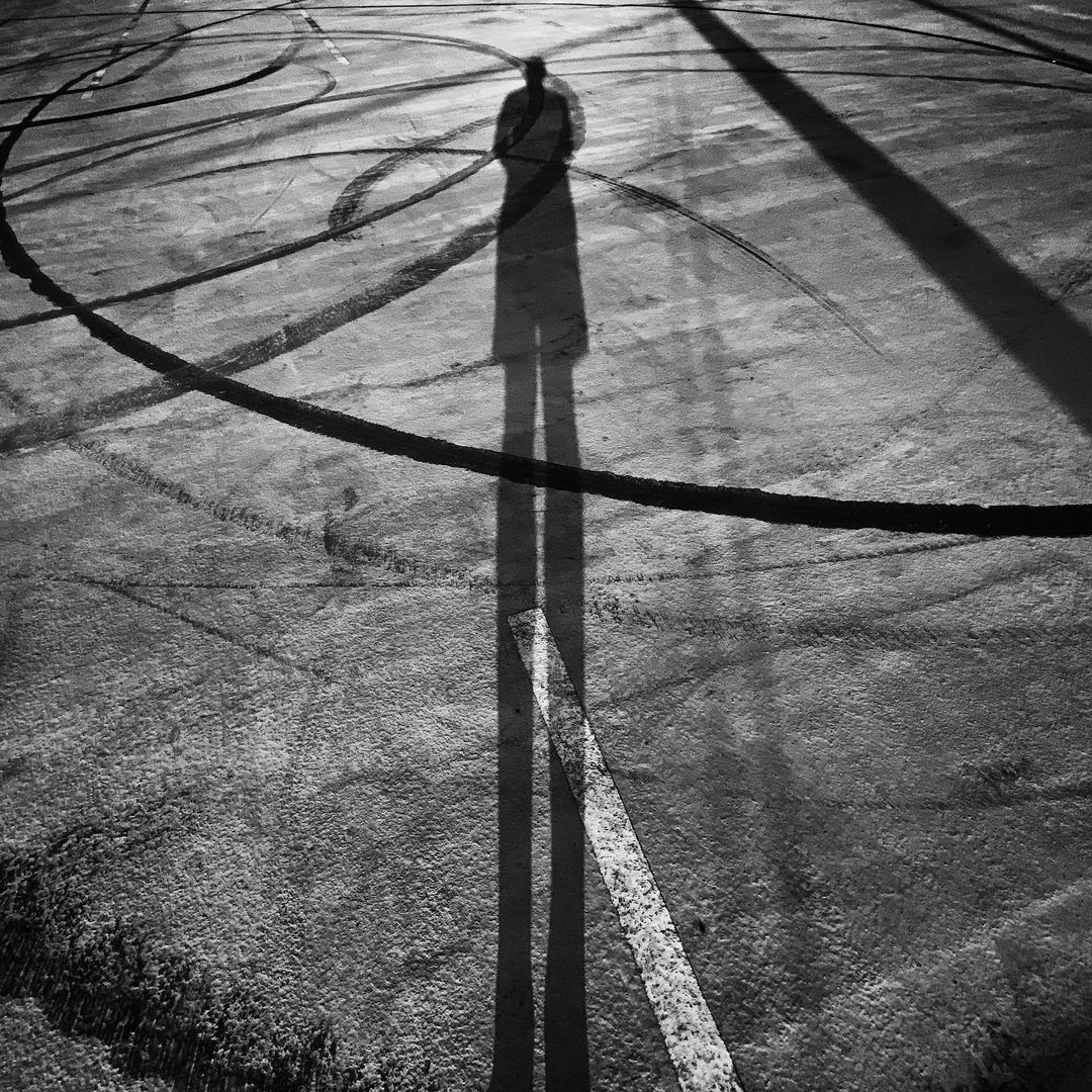 High angle black and white shot of a long shadow cast by a person on a cracked asphalt surface with curved tire marks and lines.
