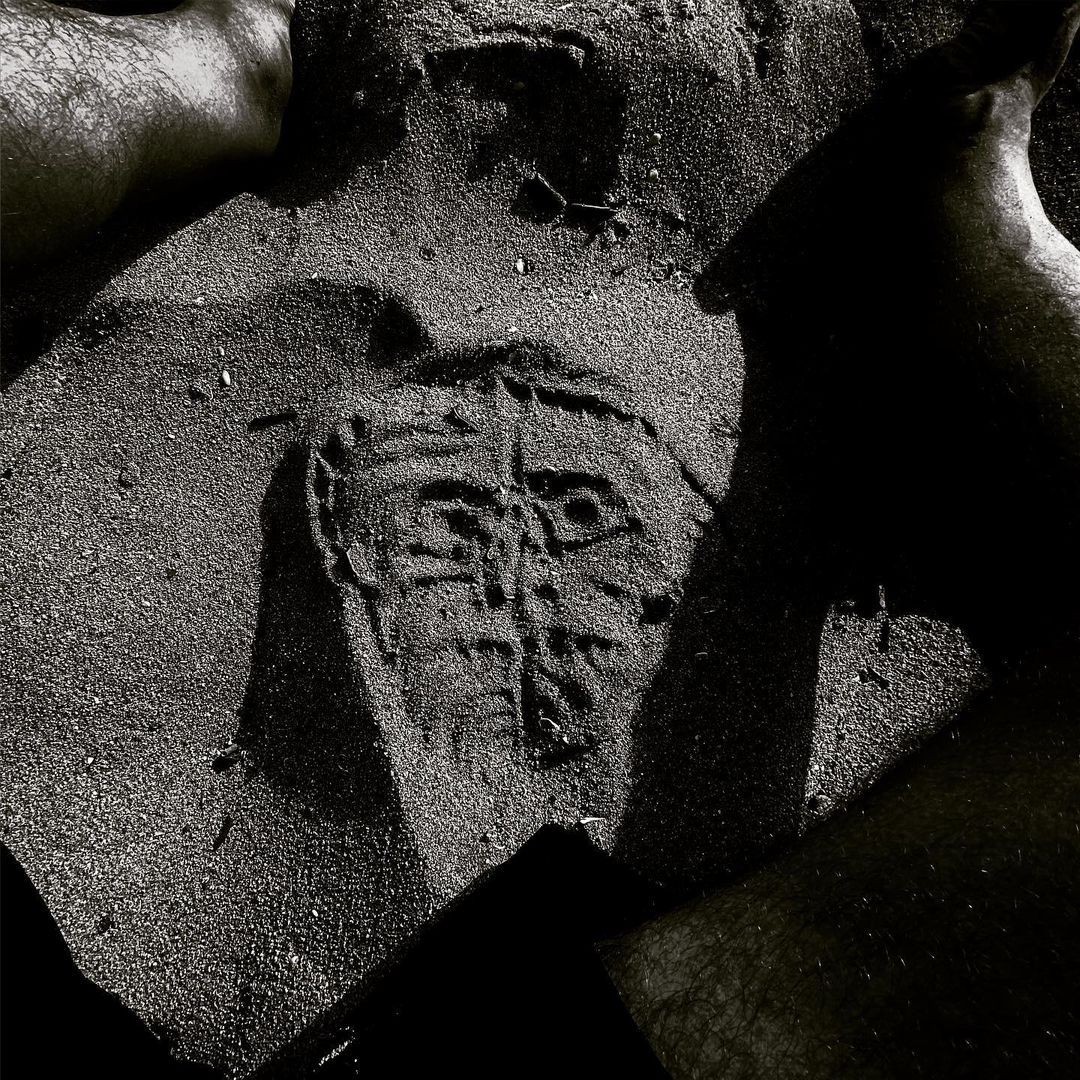 Monochrome image of a face-like design etched into sand, surrounded by partial views of four human feet; grainy texture, abstract art.