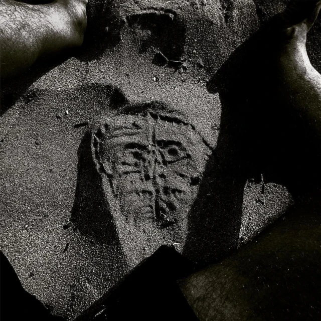 Monochrome image of a face-like design etched into sand, surrounded by partial views of four human feet; grainy texture, abstract art.