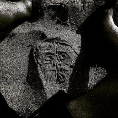 Monochrome image of a face-like design etched into sand, surrounded by partial views of four human feet; grainy texture, abstract art.