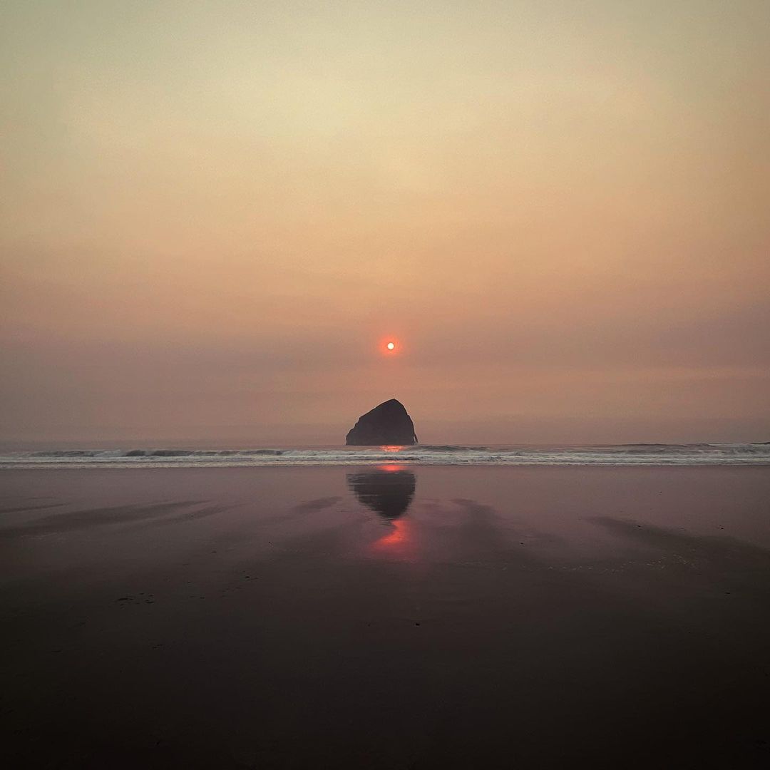Coastal image featuring a sea stack silhouetted against the sun at sunset on a beach reflecting the light and colors of the scene.