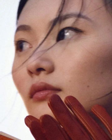 Close-up portrait of an East Asian woman with dark hair blowing across her face and gloved fingers visible in the lower foreground.