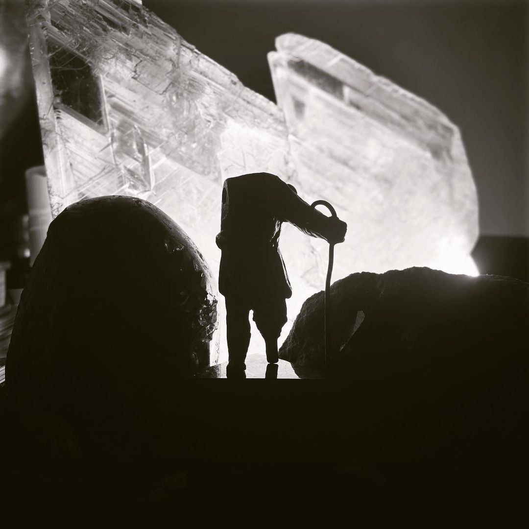 Silhouette of a person with a cane standing between two dark rocks, with a large crystal formation in the background.