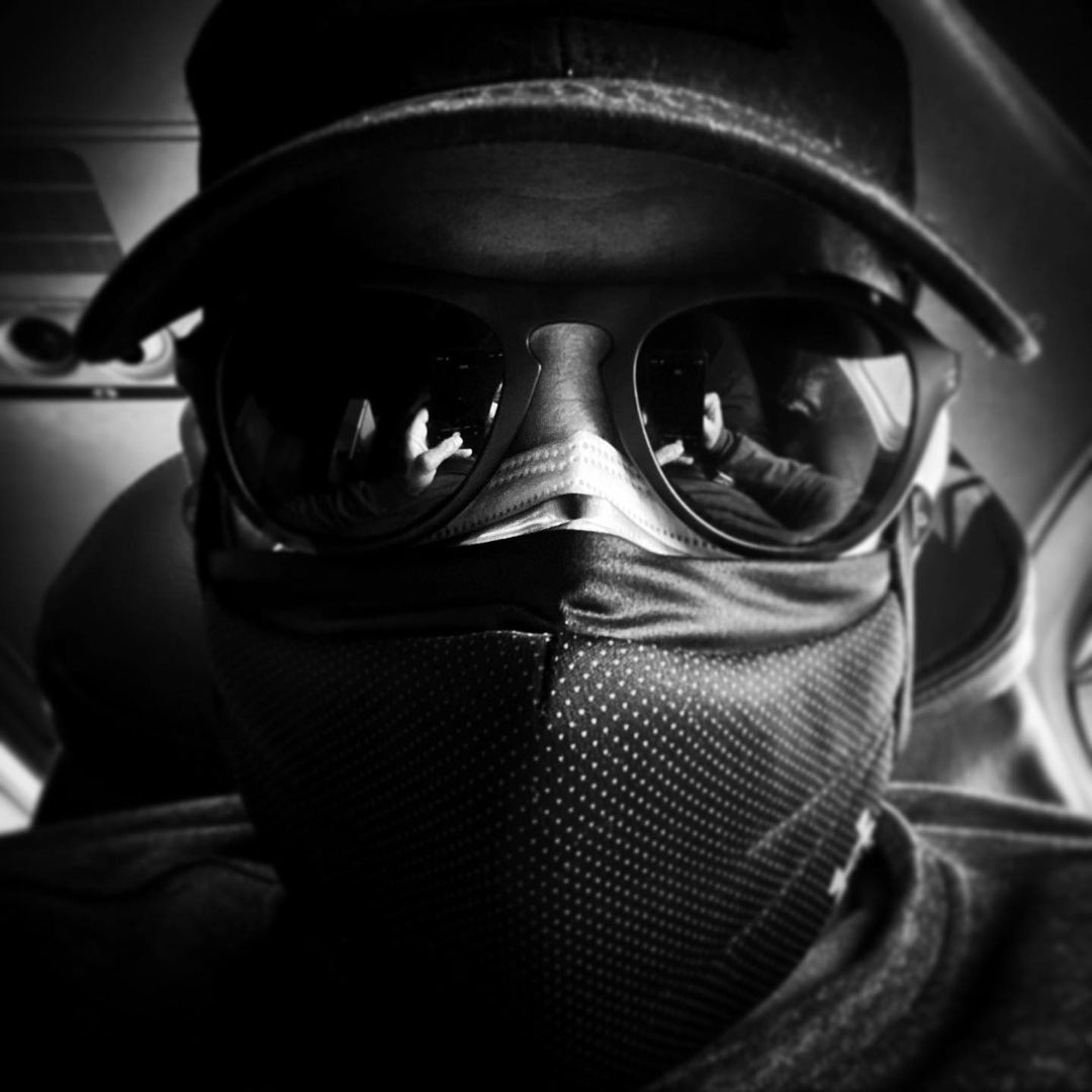 Person in hat and sunglasses, wearing a patterned face covering, poses indoors in a close-up studio-style shot, emphasizing mystery.