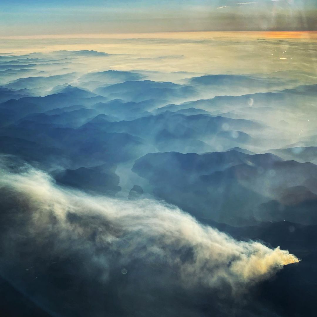 Aerial view shows layered blue mountain ranges under a hazy sky with white smoke plume rising from one peak in the foreground.