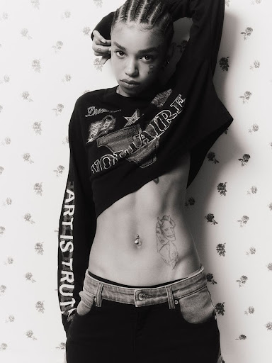 A woman with braided hair poses with a partial view of her abdomen. She wears a black long-sleeved shirt and black pants with light wash denim.