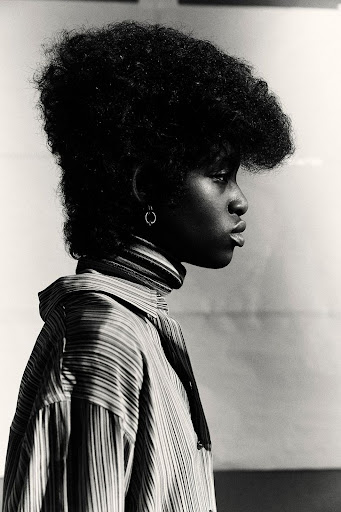 Profile portrait of a young Black woman with an afro hairstyle, wearing a striped high-neck top, against a light grey studio backdrop.
