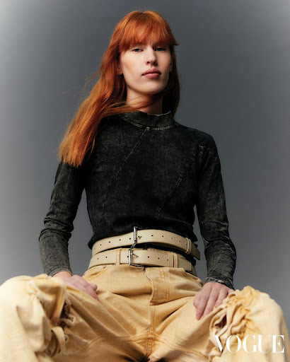 A red-haired woman is sitting, posing in a studio wearing a long-sleeve top, a double belt, and tan pants in a studio setting.