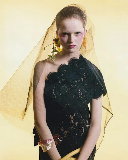 Woman in black lace dress and sheer veil with floral detail posing against a yellow backdrop, beauty and fashion editorial.