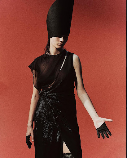 Elegant portrait of a young woman in a black dress and sculptural hat against a red background, showcasing high fashion and modern style.