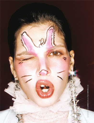 Close-up of a woman with bunny face paint winking at the camera, wearing large earrings and a white ruffled collar.