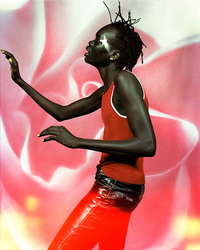 Person with painted star on face poses in front of a floral background. They wear a red tank top, leather pants and have yellow nail polish.