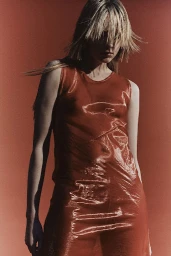 Model in a shiny reddish-orange sleeveless top against a warm terracotta background