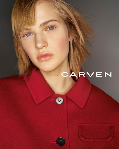 Close-up portrait of a model with short blonde hair wearing a red buttoned coat against a grey gradient background