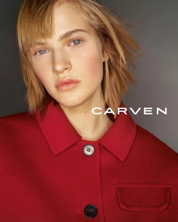 Close-up portrait of a model with short blonde hair wearing a red buttoned coat against a grey gradient background