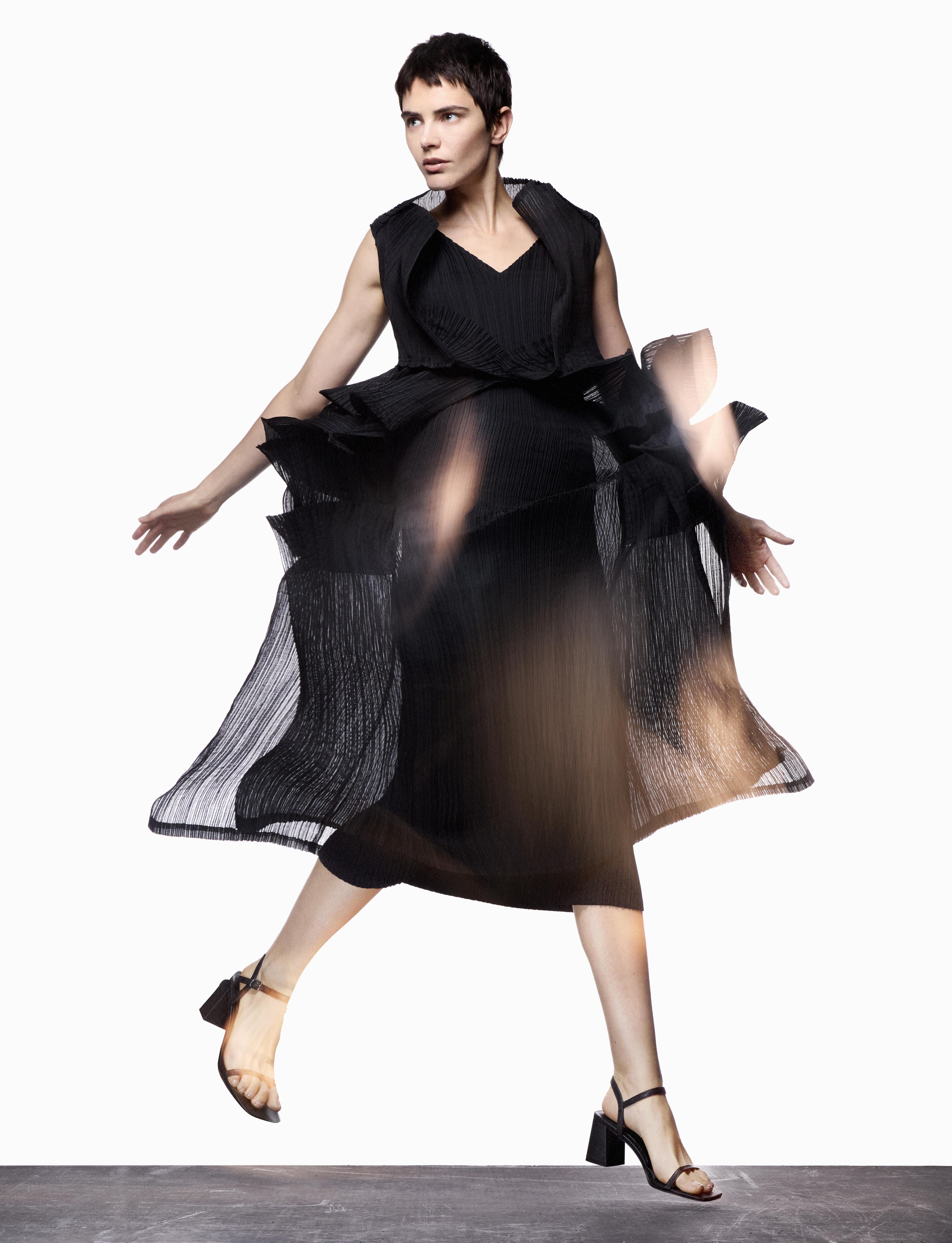 A stylish woman in a flowing black dress mid-stride against a bright white backdrop, showcasing fashion and movement with a blurred effect.
