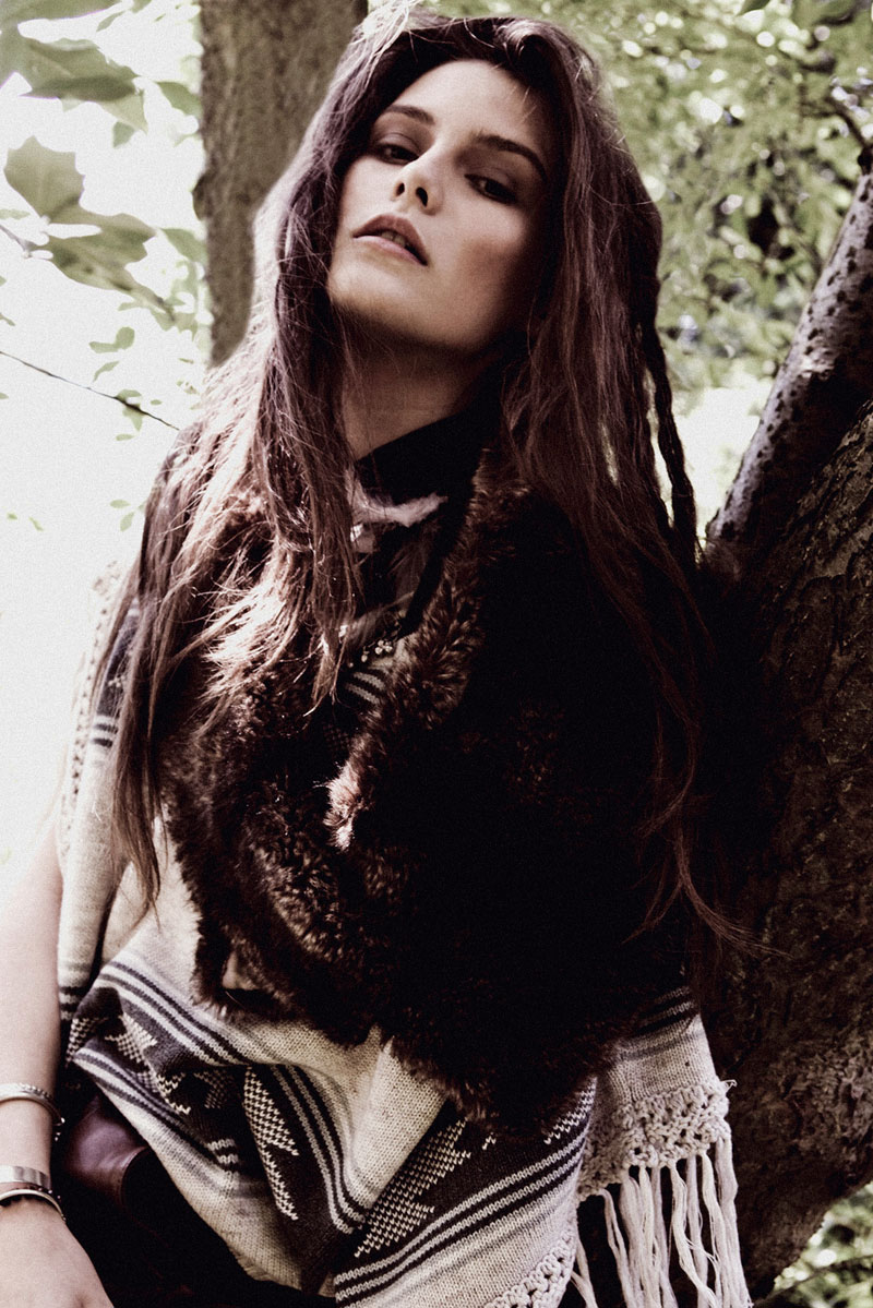 Adult woman poses in front of a tree wearing a patterned poncho with fringes and dark fur collar, soft natural lighting.