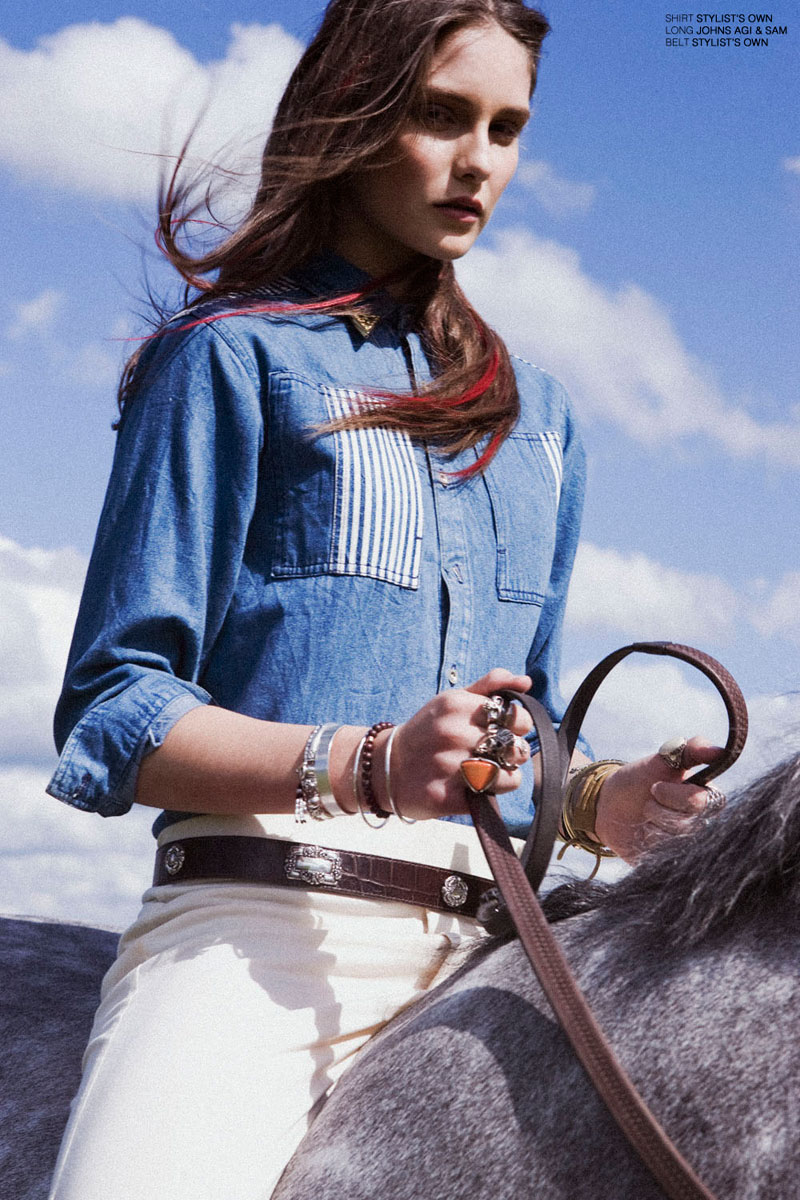 A woman wearing a denim shirt and white pants riding a grey horse with a blue sky and clouds in the background.