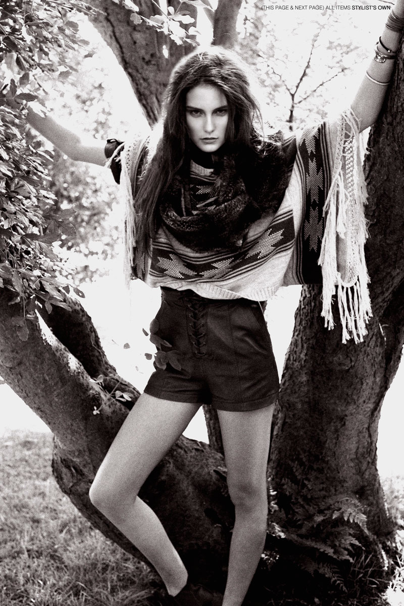 A woman stands in a black and white fashion shot, wearing shorts, a sweater, and a patterned poncho, posing with arms outstretched by a tree.