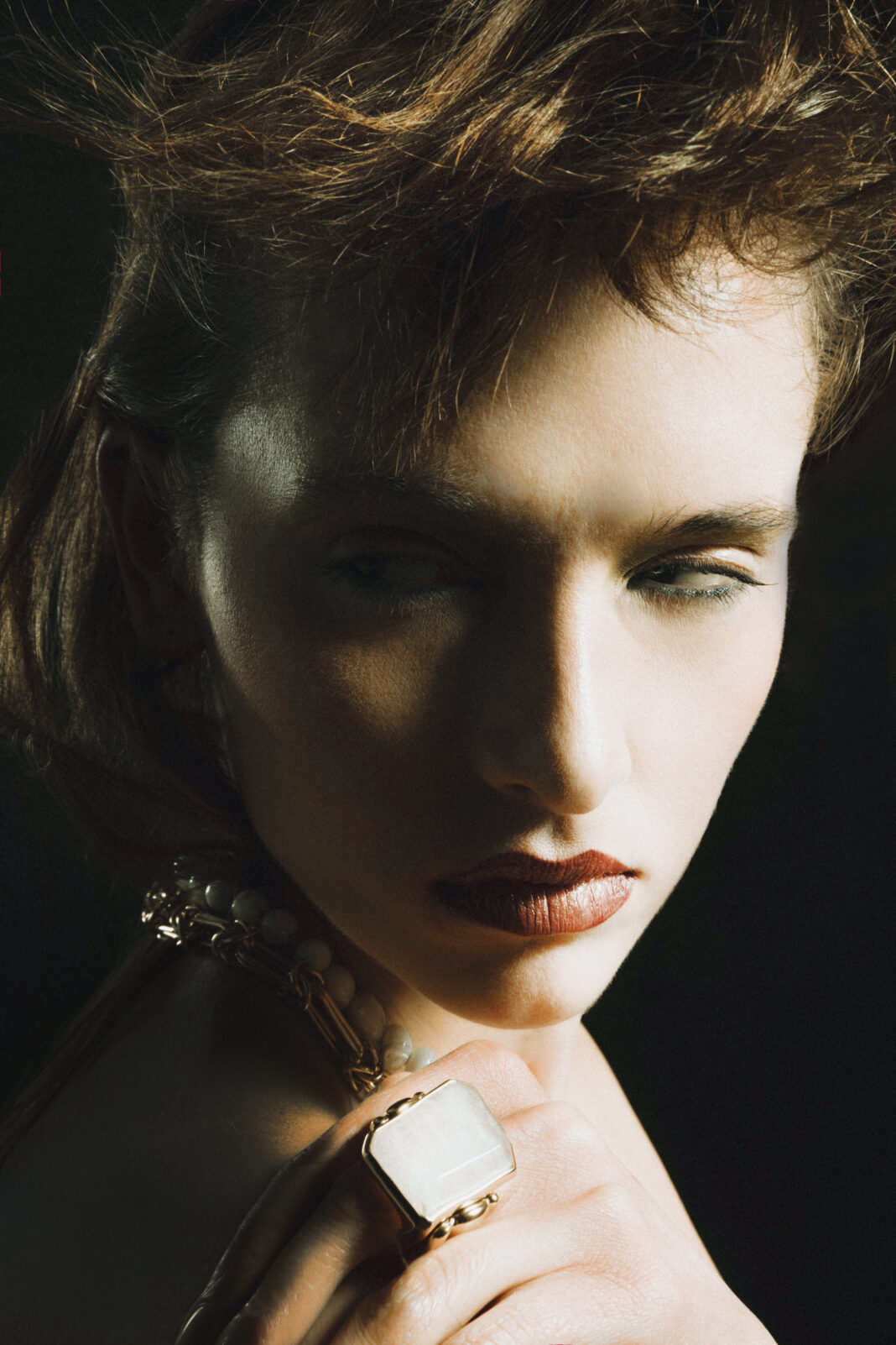 Dramatic close-up portrait of an adult female with tousled brown hair, wearing a large ring and a necklace, set against a dark backdrop.
