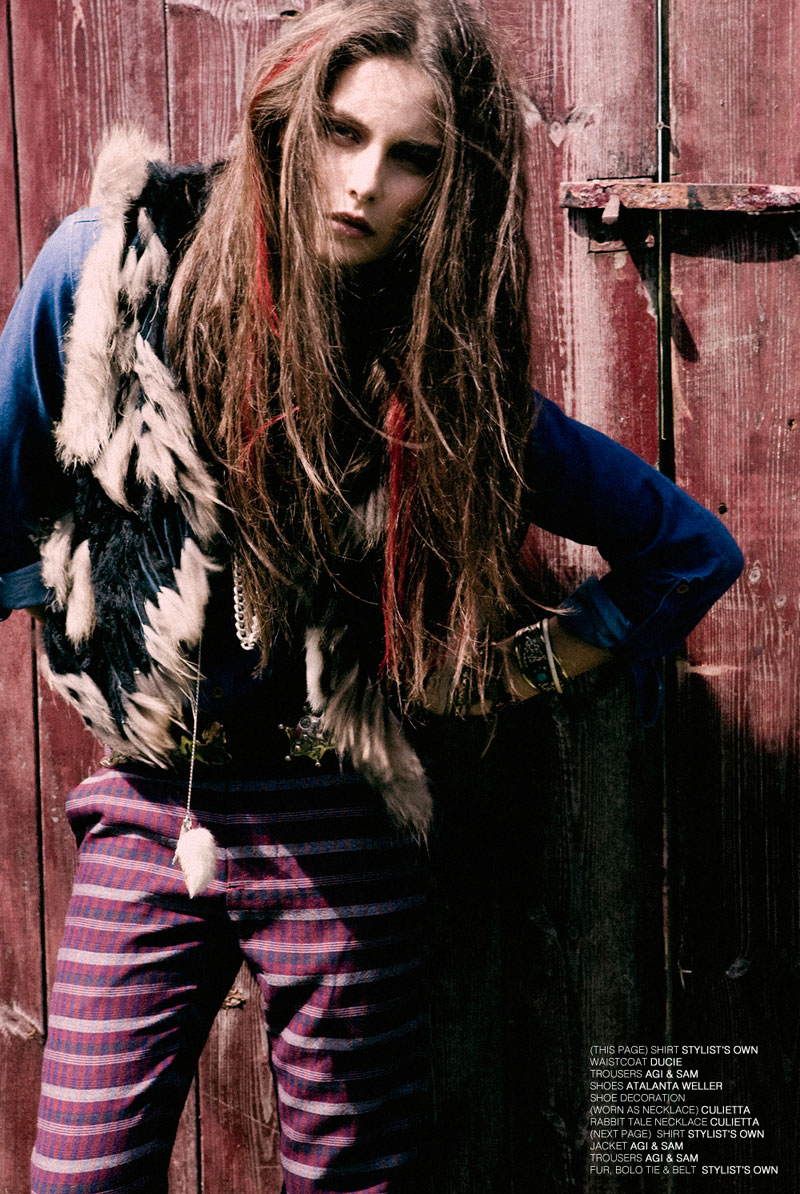 A model poses in front of a rustic wooden door, wearing a fur vest, blue top, and striped pants, styled with layered jewelry.